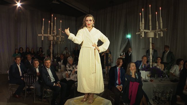 © ORF / Seitenblicke Eine Dame steht erhöht in einem Bademantel. Um sie herum sitzen Menschen an runden Tischen und schauen sie an.