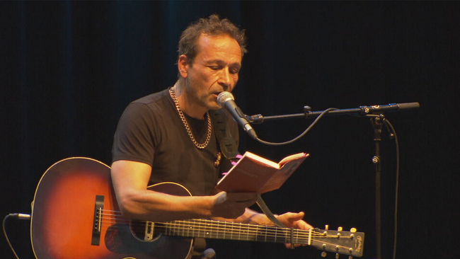 Gerald Votava sitzt mit Gitarre auf der Bühne und schaut in ein Buch von Christine Nöstlinger