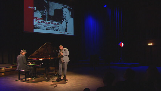 © ORF / Seitenblicke Musiker stehen auf Bühne des RKH, dahinter ist die Anzeige des Dokfilms "Jazz in Österreich" zu sehen