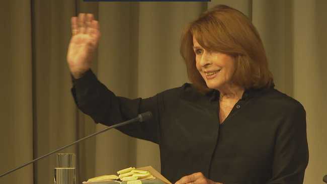 Senta Berger sitzt auf Bühne, winkt ins Publikum und lächelt. Vor ihr ein Tisch mit Büchern, aus denen sie liest
