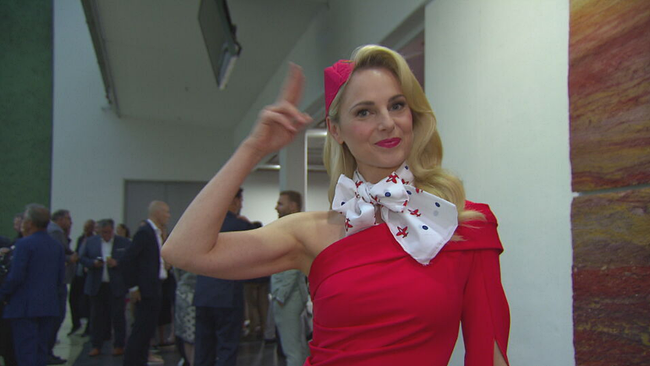 Moderatorin Silvia Schneider in rotem Stuardess-Kleid lächelt in die Kamera und macht den Piloten-Gruß 