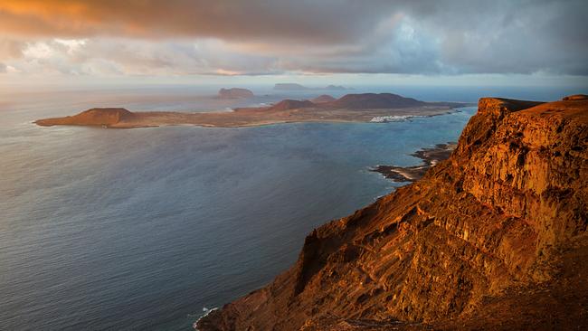 im Vordergrund schroffe, felsige Küste die rötlich vom Abendlicht beleuchtet ist. Im Meer ist im Hintergrund eine Insel zu sehen. 