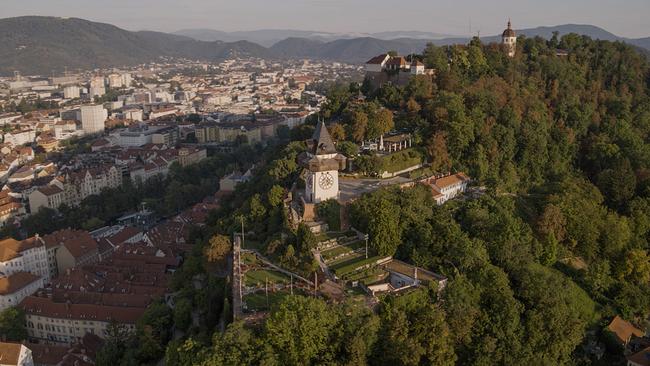Links der Bilddiagonale die Stadt Graz, in der Mitte der Grazer Uhrturm von oben. Rechts die grüne Vegetation des Schlossbergs.
