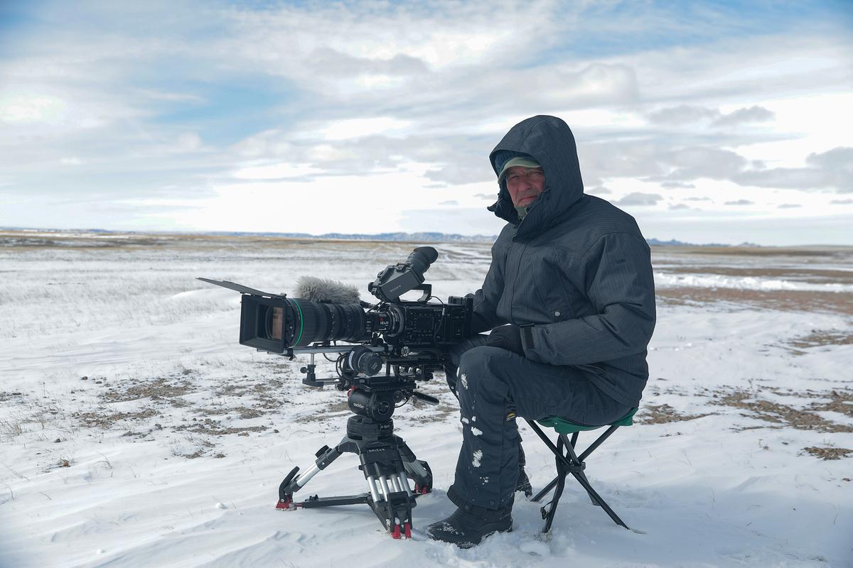 Der Regisseur Philippe Moreau sitzt dick in dunkle Kleidung eingepackt auf einem kleinen ausklappbaren Hocker, vor ihm die Kamera auf einem niedrigen Stativ. Die weite Landschaft ist schneebedeckt. 