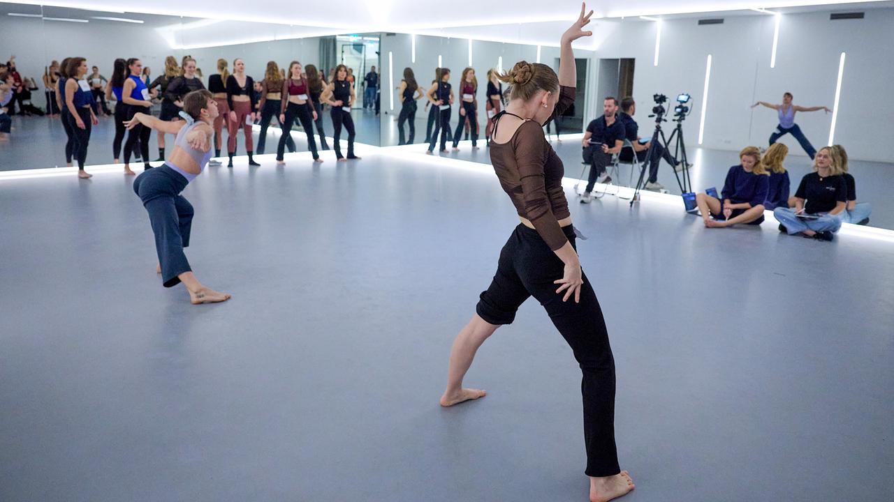 © ORF/Klaus Titzer A group of people in a modern dance studio, where two individuals in the foreground are performing a choreography while others watch or film.