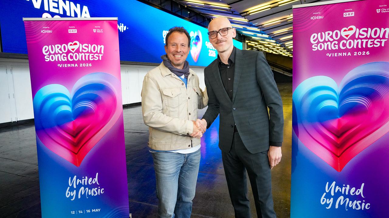 © ORF/Roman Zach-Kiesling ESC-Executive-Producer Michael Krön and Thomas Waldner (Managing Director of the Wiener Stadthalle) shake hands in front of roll-up banners of the Eurovision Song Contest 2026 in the modern, lit-up Wiener Stadthalle.