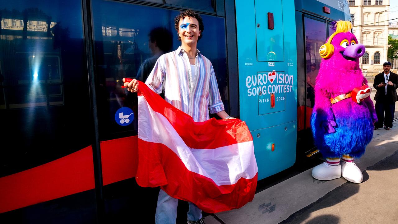COSMÓ is standing in front of the "TANZSCHEIN" special tram with Eurovision signage and holding a red-and-white-red flag, with the colorful mascot Auri in the background.