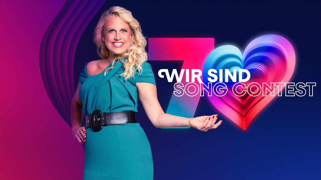 © ORF/IP Media/Peter Krivograd Barbara Schöneberger stands in front of a colorful, modernly designed logo with the number 7 and a glowing heart symbol representing the number 0 and the lettering "WIR SIND SONG CONTEST".