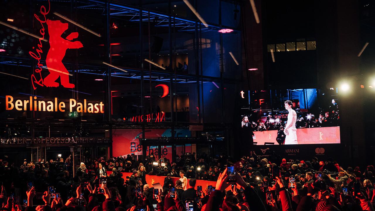 Vor dem Berlinale Palast drängen sich auf der "Berlinale 2025" zahlreiche Menschen und Fotografen an einem rot ausgeleuchteten Premierenbereich, während auf einer großen Leinwand das Geschehen vom roten Teppich gezeigt wird.