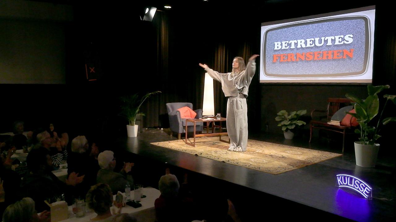 © ORF/Andreas Elsner GesmbH Kulisse Wien Malarina auf der Bühne mit ausgebreiteten Armen, im Hintergrund auf einer Leinwand der Schriftzug „BETREUTES FERNSEHEN“, davor applaudierendes Publikum.