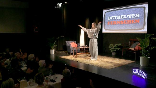 © ORF/Andreas Elsner GesmbH Kulisse Wien Malarina auf der Bühne mit ausgebreiteten Armen, im Hintergrund auf einer Leinwand der Schriftzug „BETREUTES FERNSEHEN“, davor applaudierendes Publikum.