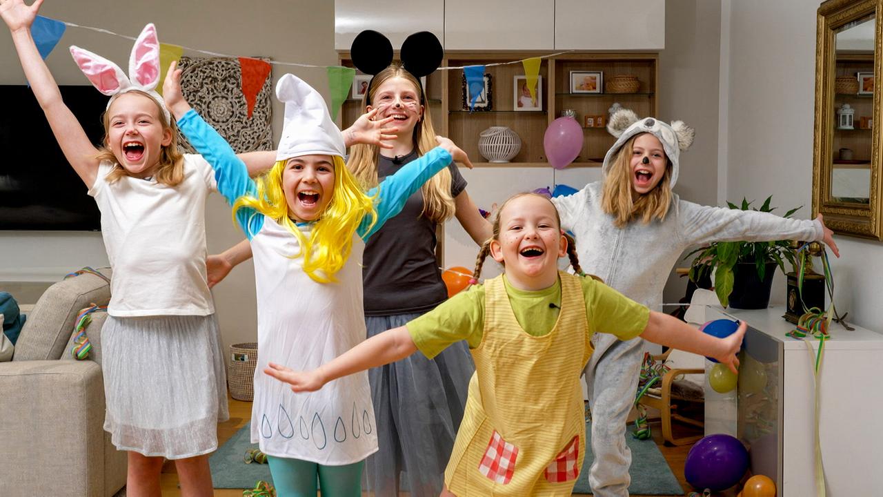 Mehrere Kinder in bunten Kostümen tanzen und posieren ausgelassen in einem festlich dekorierten Wohnzimmer mit Girlanden und Luftballons.