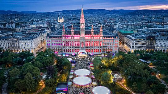 Das Wiener Rathaus ist bei Abenddämmerung festlich beleuchtet, davor füllt ein großes Event das Parkgelände.