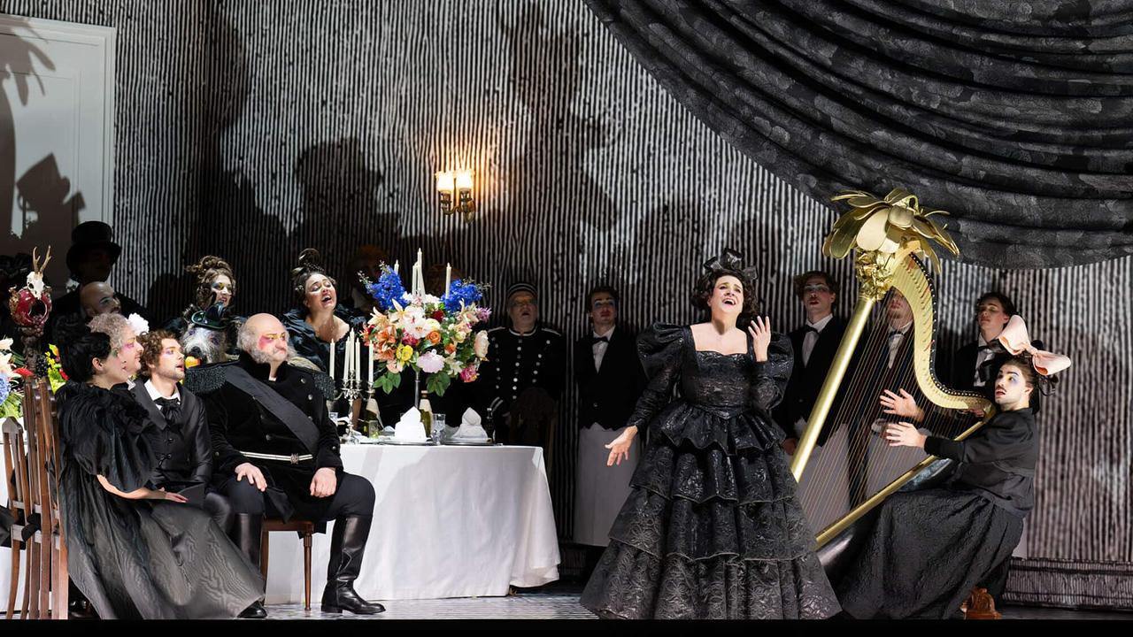 Bühnenszene aus "Il viaggio a Reims": Cecilia Bartoli als Corinna in einem wallenden schwarzen Kleid singt vor einem festlich gedeckten Tisch, umgeben von einem Harfe-Spieler und weiteren Darstellern.