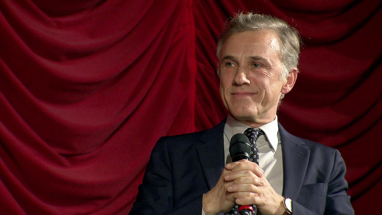 © ORF/Kurt Mayer Film/Viennale Film Festival Christoph Waltz beim Interview mit Gabi Flossmann, im Anzug, Hemd und Krawatte, mit dem Mikrofon in der Hand erwartet er beim Interview freundlich lächelnd die nächste Frage