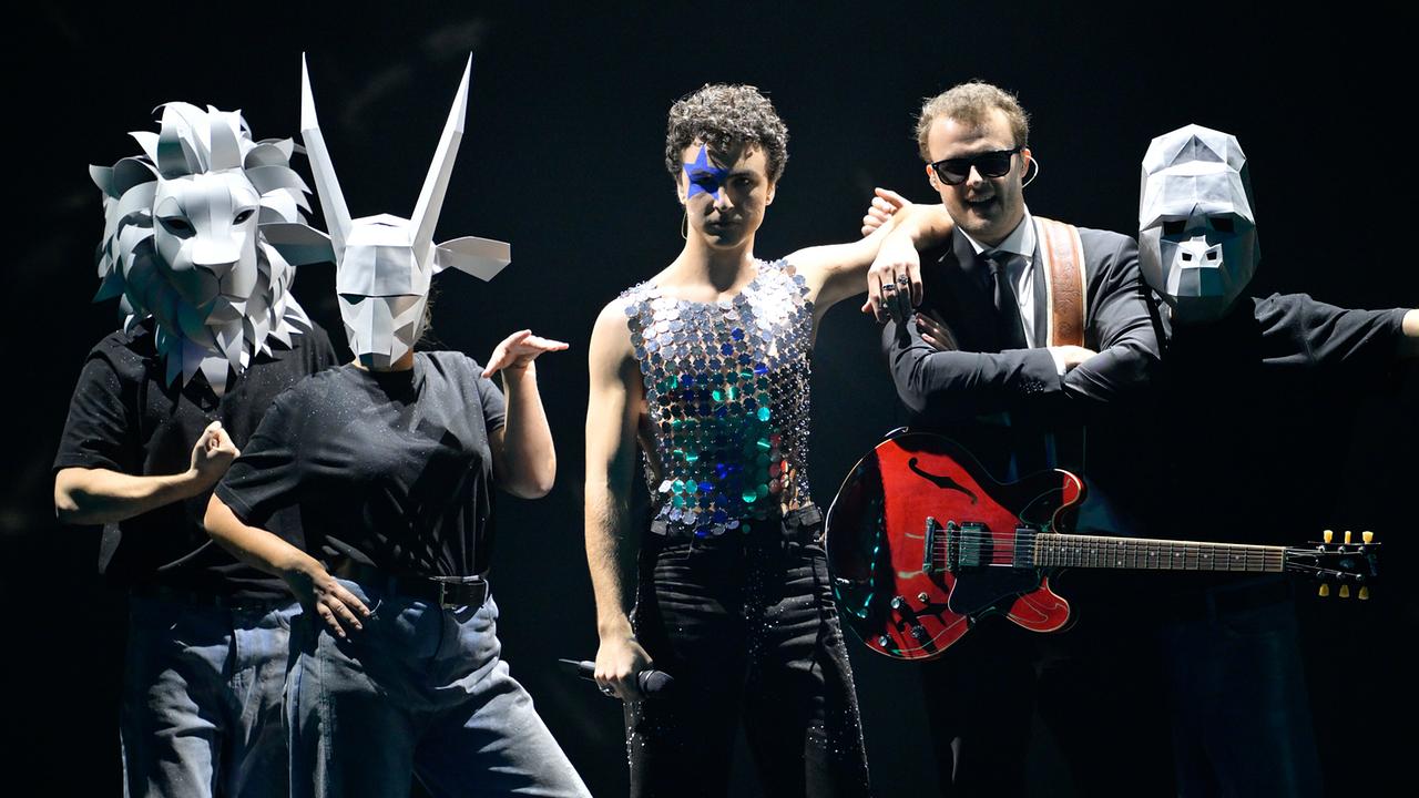 © ORF/Thomas Ramstorfer Mehrere Performer stehen auf einer dunklen Bühne, einige mit kunstvollen Tiermasken, COSMÓ in der Mitte mit einem glitzernden Oberteil mit Mikrofon