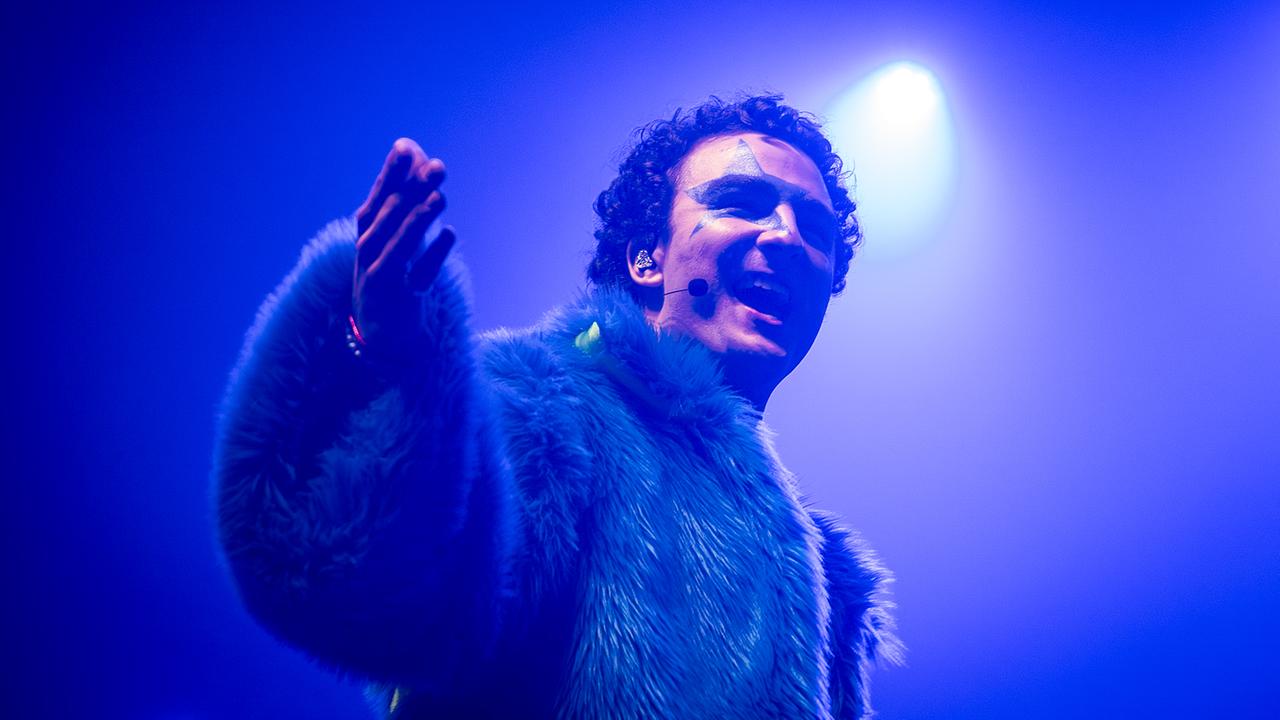Eurovision in Concert - Amsterdam: COSMÓ in a fluffy blue faux fur jacket stands in the stage light and stretches out a hand, surrounded by intense blue-violet spotlight.