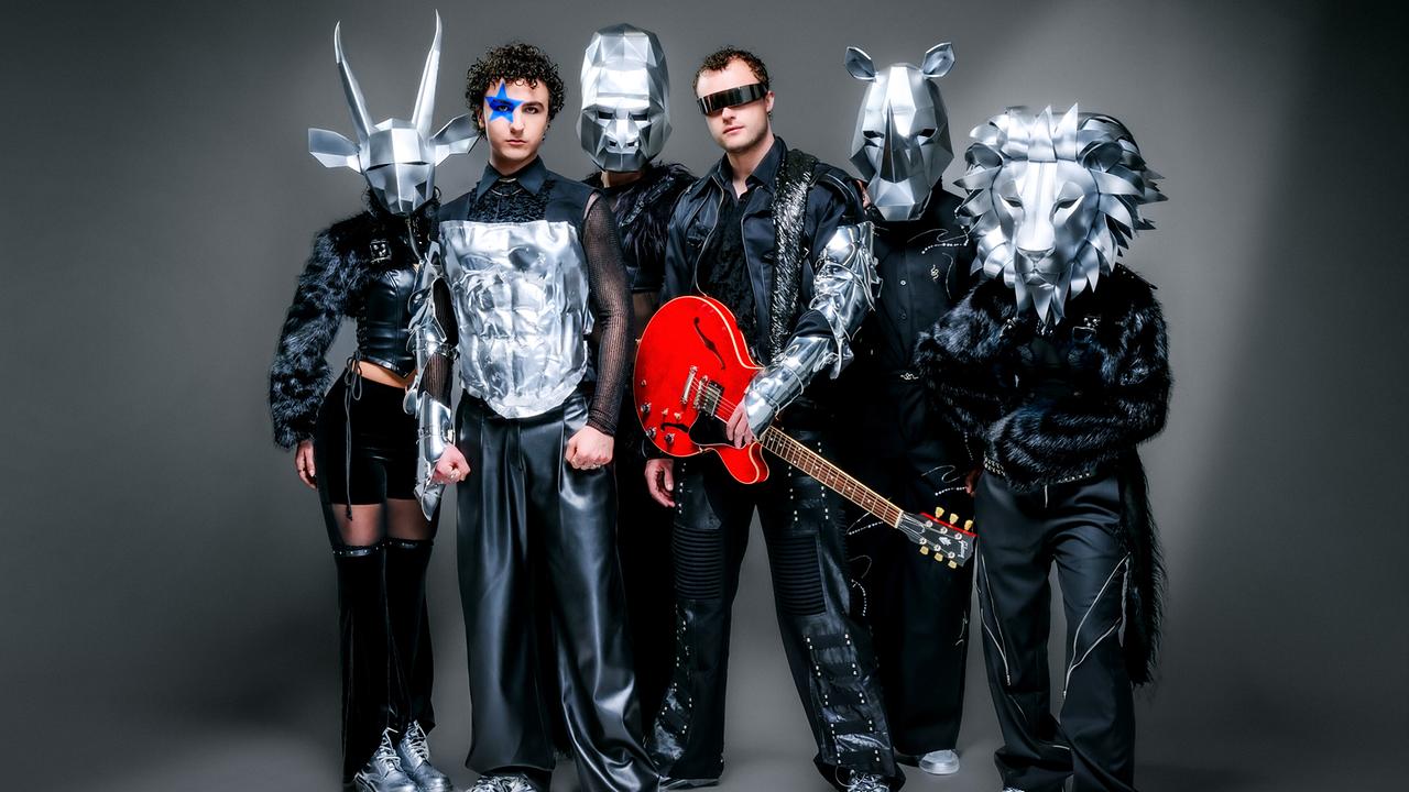 COSMÓ mit blauem Stern im Gesicht und fünf weitere Personen in futuristischen, schwarzen Outfits mit silbernen Tiermasken posieren vor neutralem Hintergrund, eine davon hält eine rote E‑Gitarre.