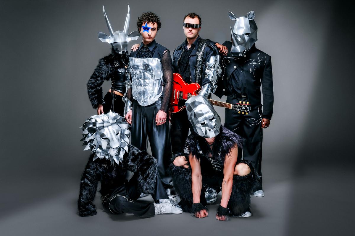 COSMÓ mit blauem Stern im Gesicht und fünf weitere Personen in futuristischen, schwarzen Outfits mit silbernen Tiermasken posieren vor neutralem Hintergrund, eine davon hält eine rote E‑Gitarre.