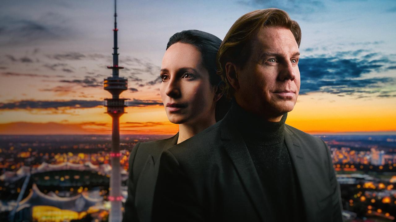 Stephanie Stumph und Thomas Heinze stehen Rücken an Rücken im Vordergrund, dahinter ist die Stadtlandschaft von München mit Fernsehturm bei Sonnenuntergang zu sehen.
