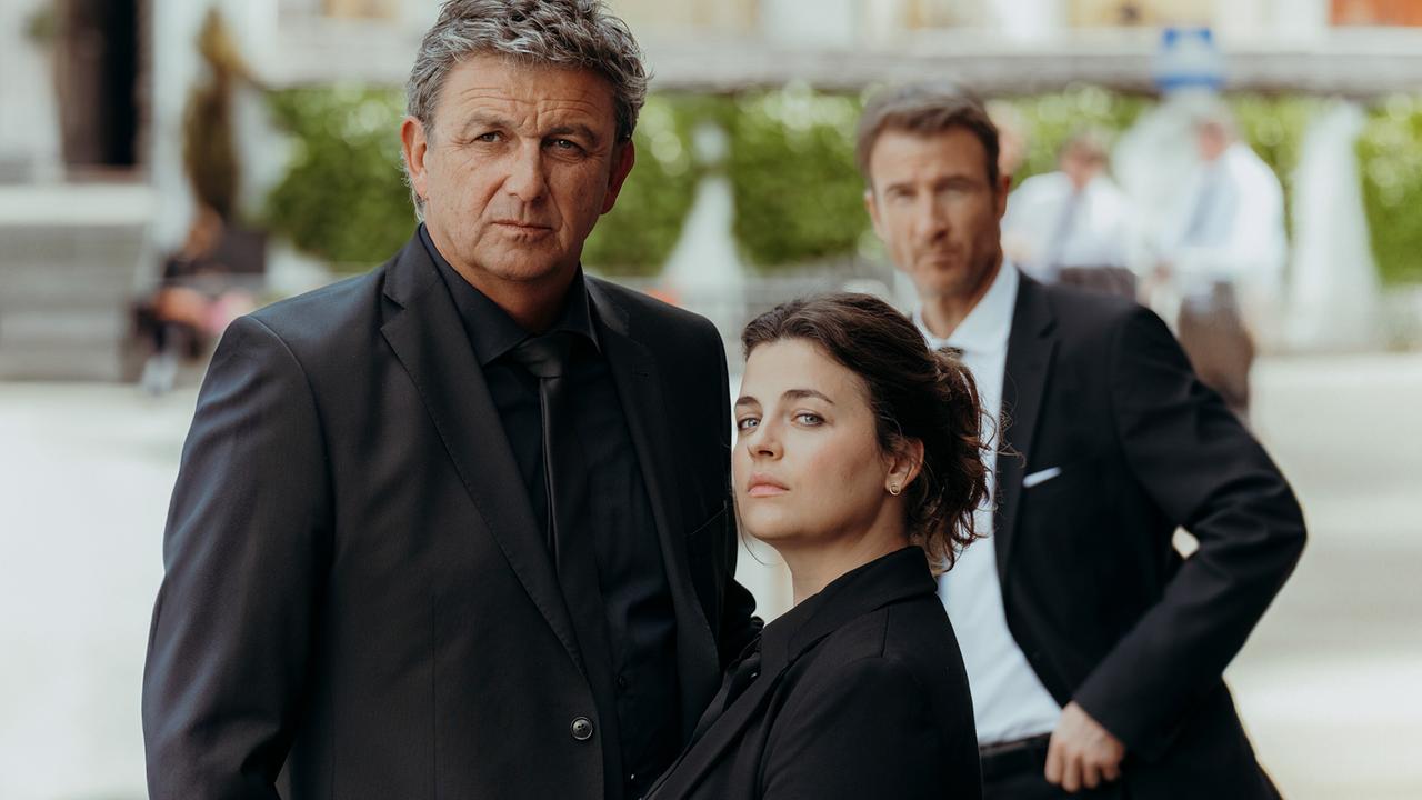 Nach dem plötzlichen Tod von Rolf Pflüger: Martin und Lilli Gruber, schwarz gekleidet, stehen nebeneinander, Im Hintergrund steht Hans Gruber. Lilli ist fest entschlossen, seinem letzten Willen nachzukommen, Martin Gruber ist skeptisch.