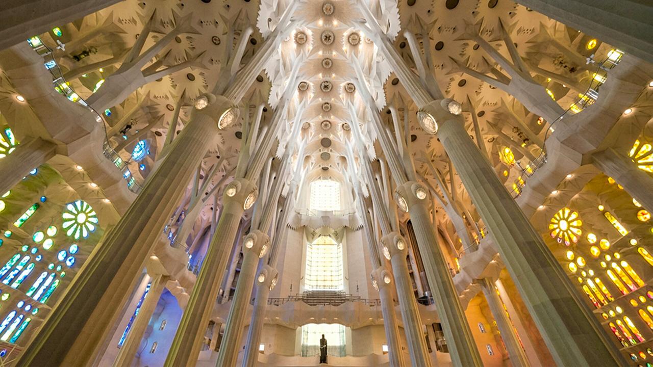 Das Bild zeigt den monumentalen Innenraum der Sagrada Família mit hoch aufragenden, verzweigten Säulen, einer kunstvollen Decke und farbigem Licht, das durch die Glasfenster einfällt.