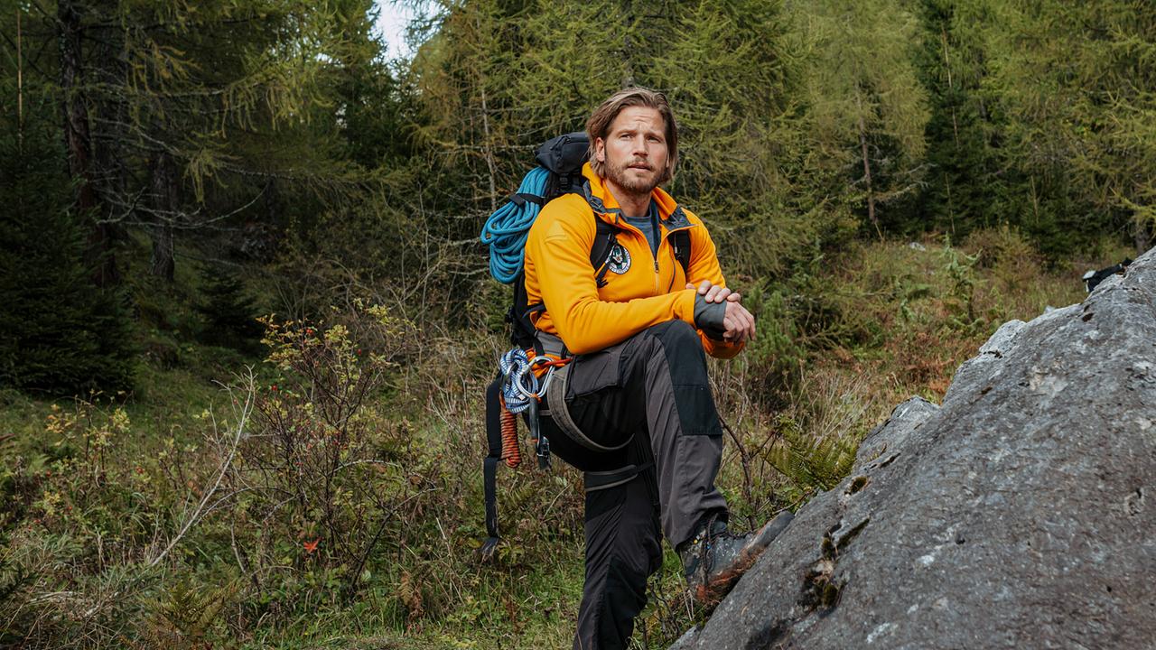 © ORF/ZDF/Sabine Finger Bergretter Markus Kofler in gelber Outdoorjacke steht mit Kletterausrüstung an einem Felsen im Wald.
