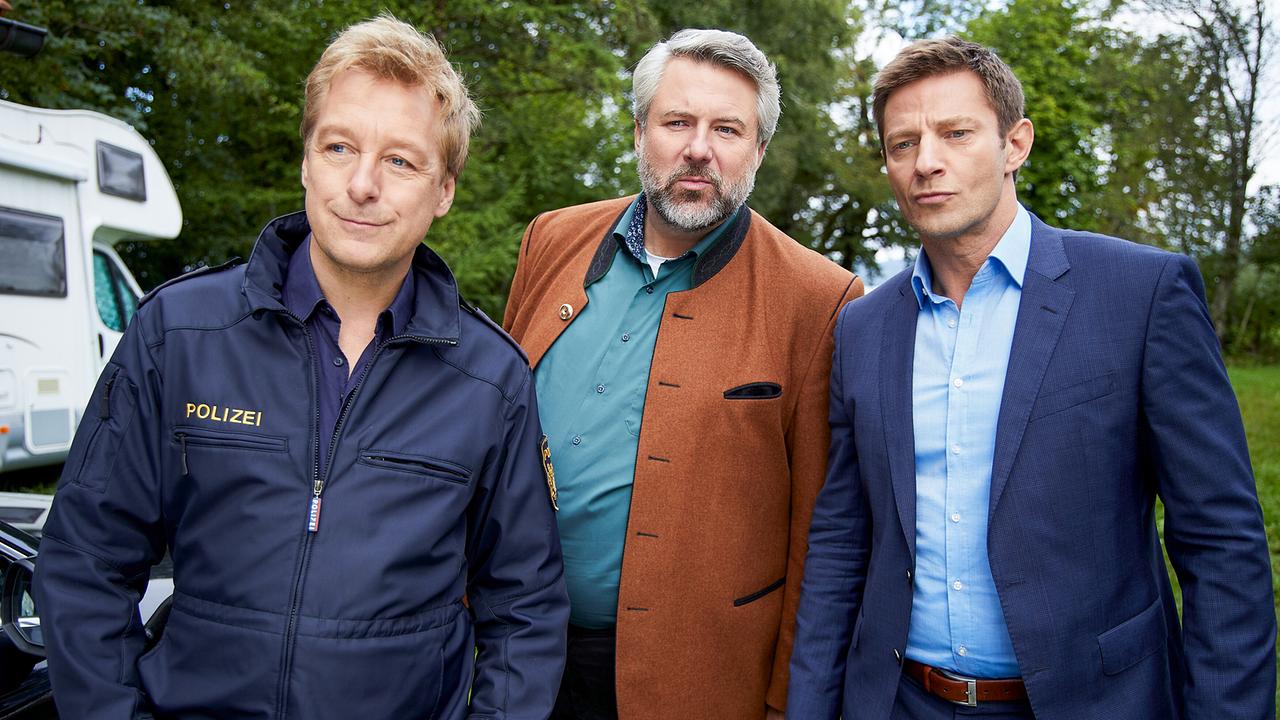 Polizeihauptmeister Mohr, Kommissar Anton Stadler und Kommissar Sven Hansen stehen nebeneinander auf einer Wiese mit einem Camper und Bäumen im Hintergrund.