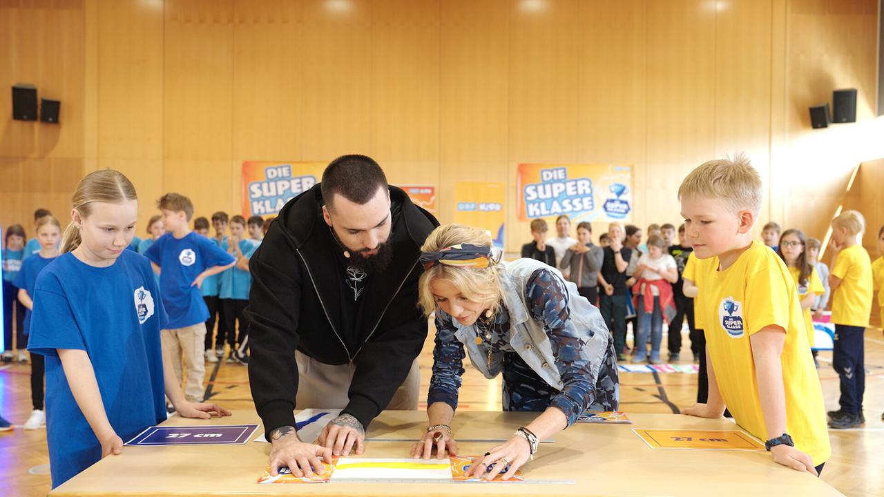 © ORF/ungefiltert Stefan und Fanny umgeben von Schüler:innen in einer Halle überprüfen, ob Magdalena oder Andreas beim Schätzspiel die Nase vorne hat.