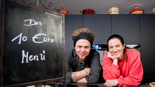 Lisa Gadenstätter mit Spitzenkoch Christoph ‚Krauli‘ Held lehnen in einer Küche an der Arbeitsfläche; daneben steht eine Tafel mit der Aufschrift „Das 10 Euro Menü“.