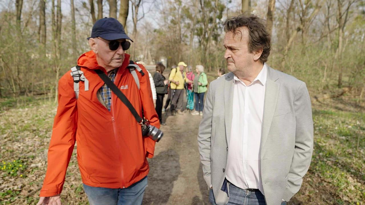 Beim Seniorenwandertag gehen Hanno Settele im Gespräch mit Pensionist Erich Kdolsky auf einem Waldweg nebeneinander, während im Hintergrund eine kleine Gruppe von Menschen folgt.