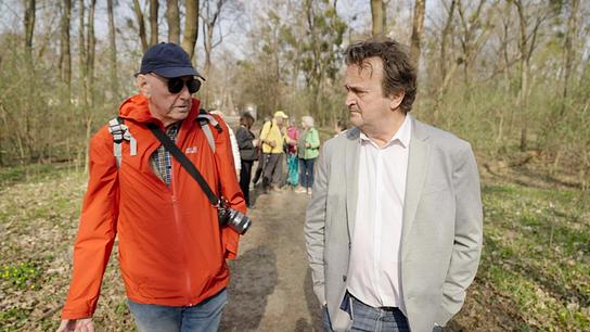 Beim Seniorenwandertag gehen Hanno Settele im Gespräch mit Pensionist Erich Kdolsky auf einem Waldweg nebeneinander, während im Hintergrund eine kleine Gruppe von Menschen folgt.