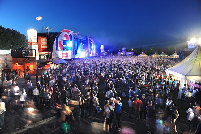 Eine riesige Menschenmenge abends beim Donauinselfest, die vor einer hell beleuchteten Bühne unter freiem Himmel feiert.