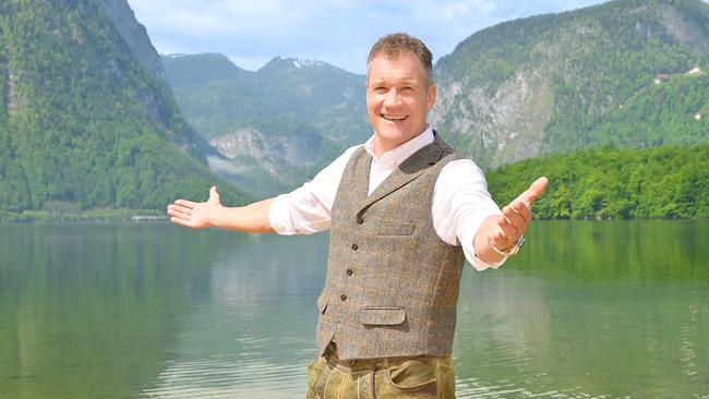 © ORF/Thomas Ramstorfer Armin Assinger in traditioneller Tracht steht mit ausgebreiteten Armen auf einem Holzsteg an einem alpinen See, umgeben von Bergen und grünen Hängen.