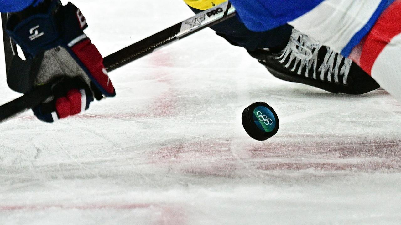 © APA/AFP/JULIEN DE ROSA Eine Detailaufnahme zeigt einen Eishockeyschläger und Schlittschuhe im Spielmoment, während ein Puck mit olympischem Logo über das Eis gleitet im Rahmen der Olympischen Winterspiele 2026.