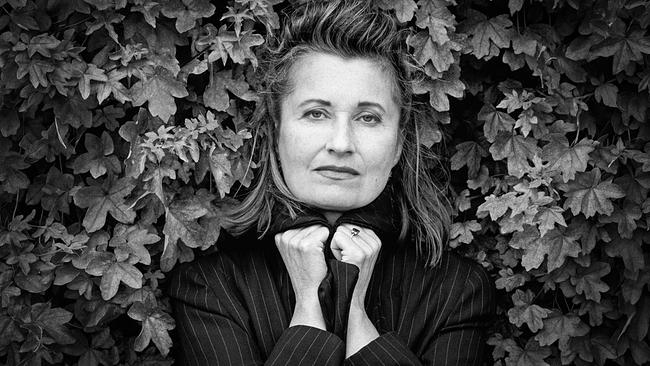 Elfriede Jelinek blickt in die Kamera und hält den Kragen ihrer dunklen, gestreiften Jacke vor dichtem Laub fest.