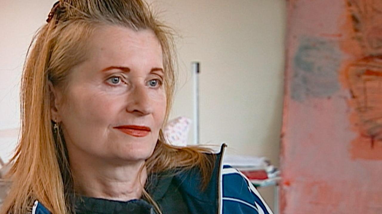 © ORF/Polyfilm Nahaufnahme von Elfriede Jelinek mit rot-blonden Haaren und blauer Jacke in einem Innenraum neben einem abstrakten Gemälde.