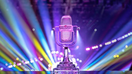 Die ESC 2026 Trophy in Mikrofonform steht vor einer Bühne mit bunten Lichtstrahlen und trägt das Logo des Eurovision Song Contest.