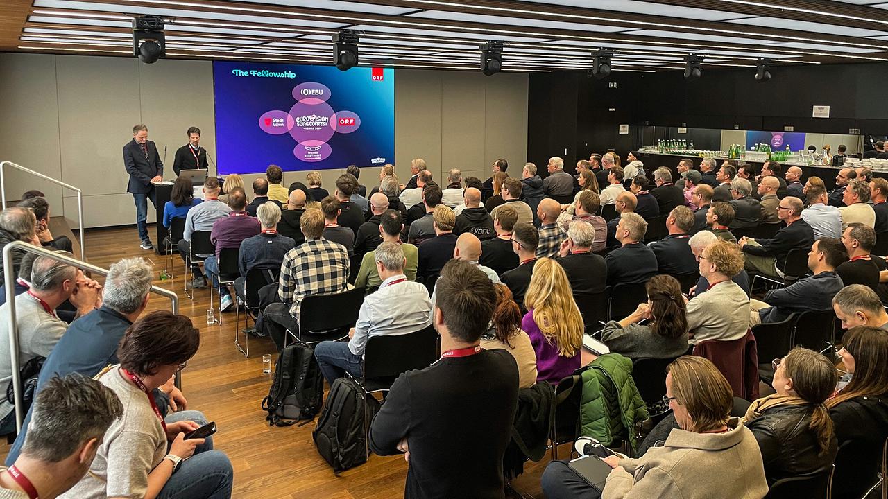 © ORF/Benedikt Fuchs A large group of people in a modern conference room following a lecture by two presenters at the ESC Production Meeting in front of a screen.