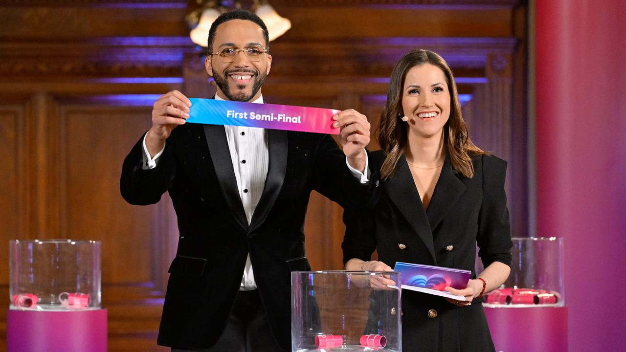 © ORF/Thomas Ramstorfer Cesár Sampson presents a colorful sign with the inscription "First Semi-Final" alongside Alexandra Maritza Wachter at a drawing in a festively decorated room.