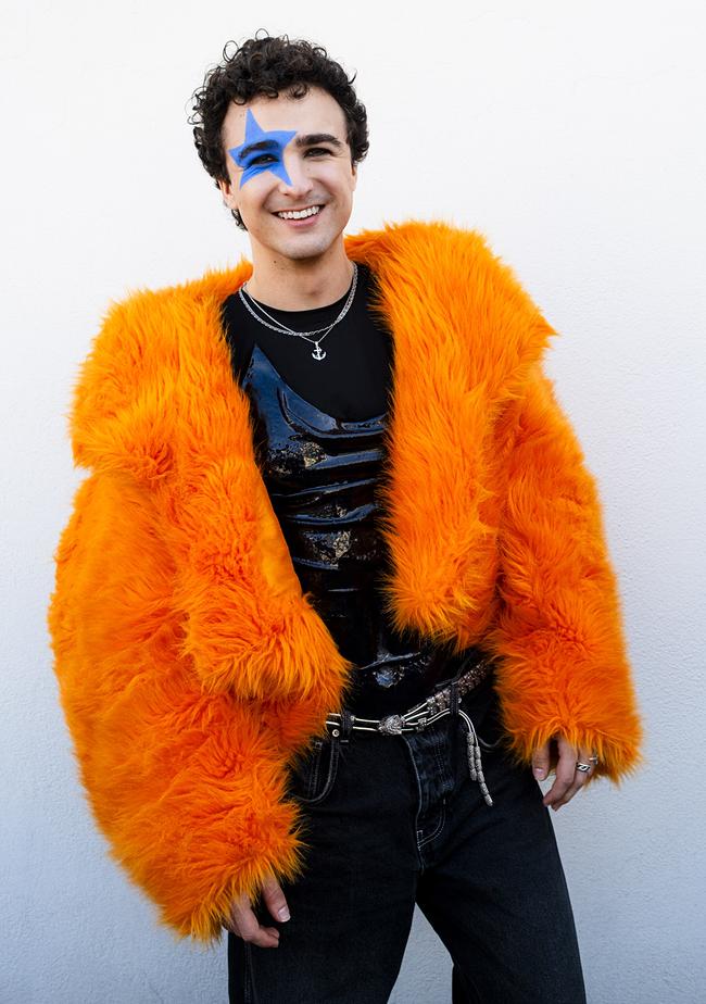 © ORF/toechterundsoehne/Matthias Heschl COSMÓ trägt eine auffällige orangefarbene Felljacke über einem schwarzen Outfit und posiert selbstbewusst vor einer schlichten weißen Wand.