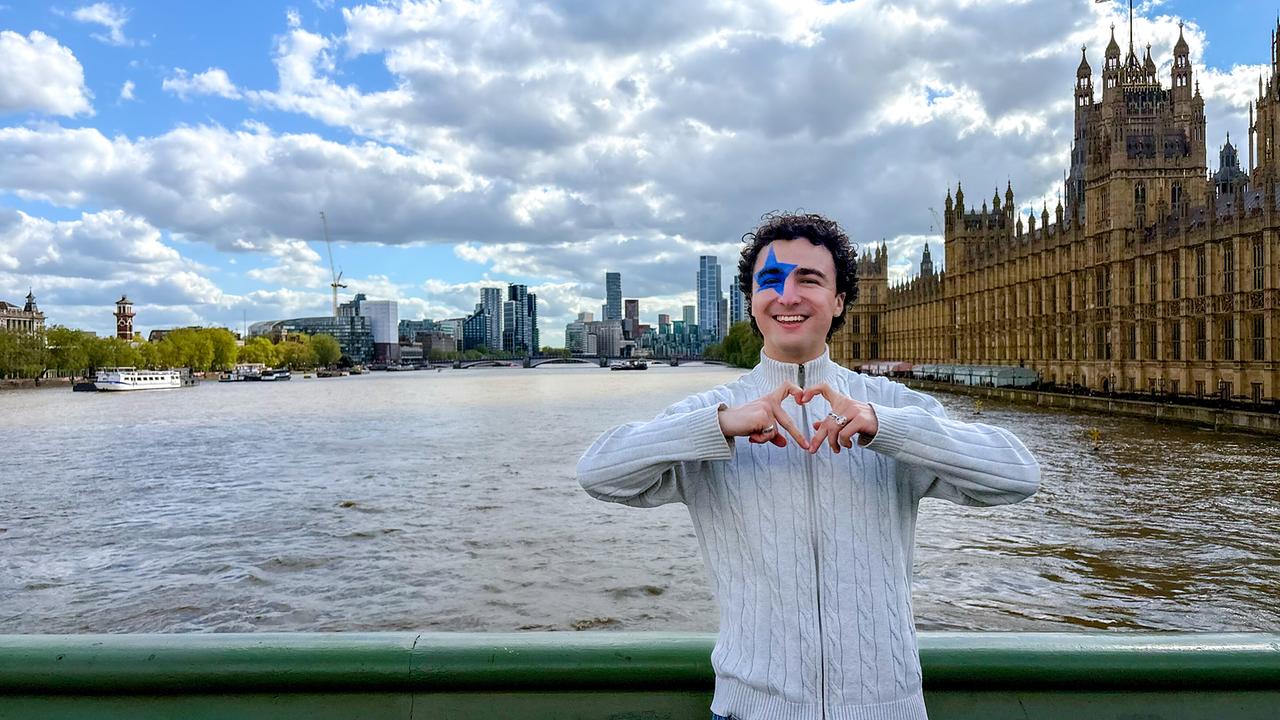 COSMÓ stands on a bridge over the Thames forming a heart with his hands, with London landmarks visible in the background.