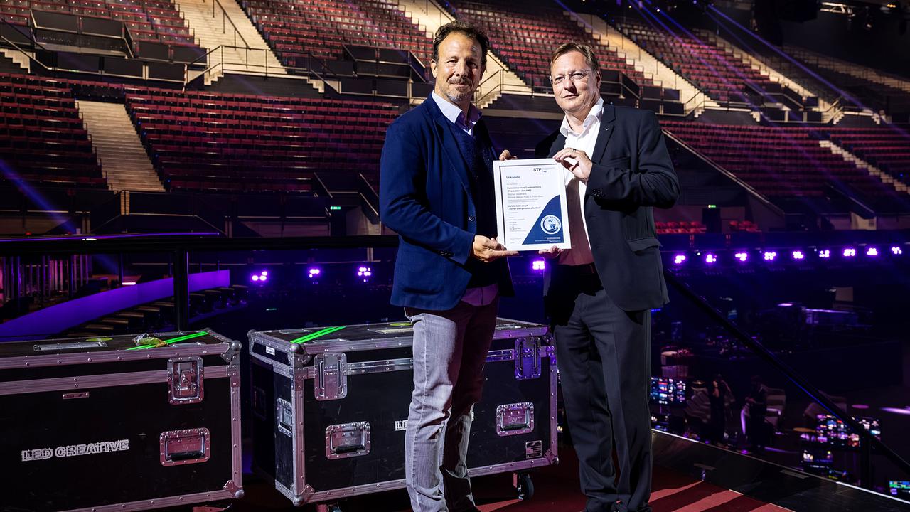 Michael Krön (Executive Producer des Eurovision Song Contest) and Roland Pichler (Deputy Director General of the AUVA) stand on a stage in a large, empty event hall and together hold the quality seal, surrounded by technical equipment and illuminated seating.
