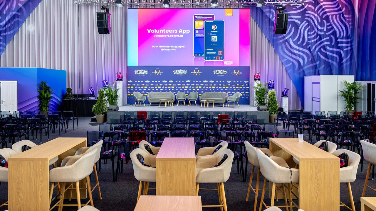 The Media Center in the Wiener Stadthalle: A large event hall with a stage, seating and lounge areas is prepared for an event; a presentation about the "Volunteers App" is being shown on the stage.