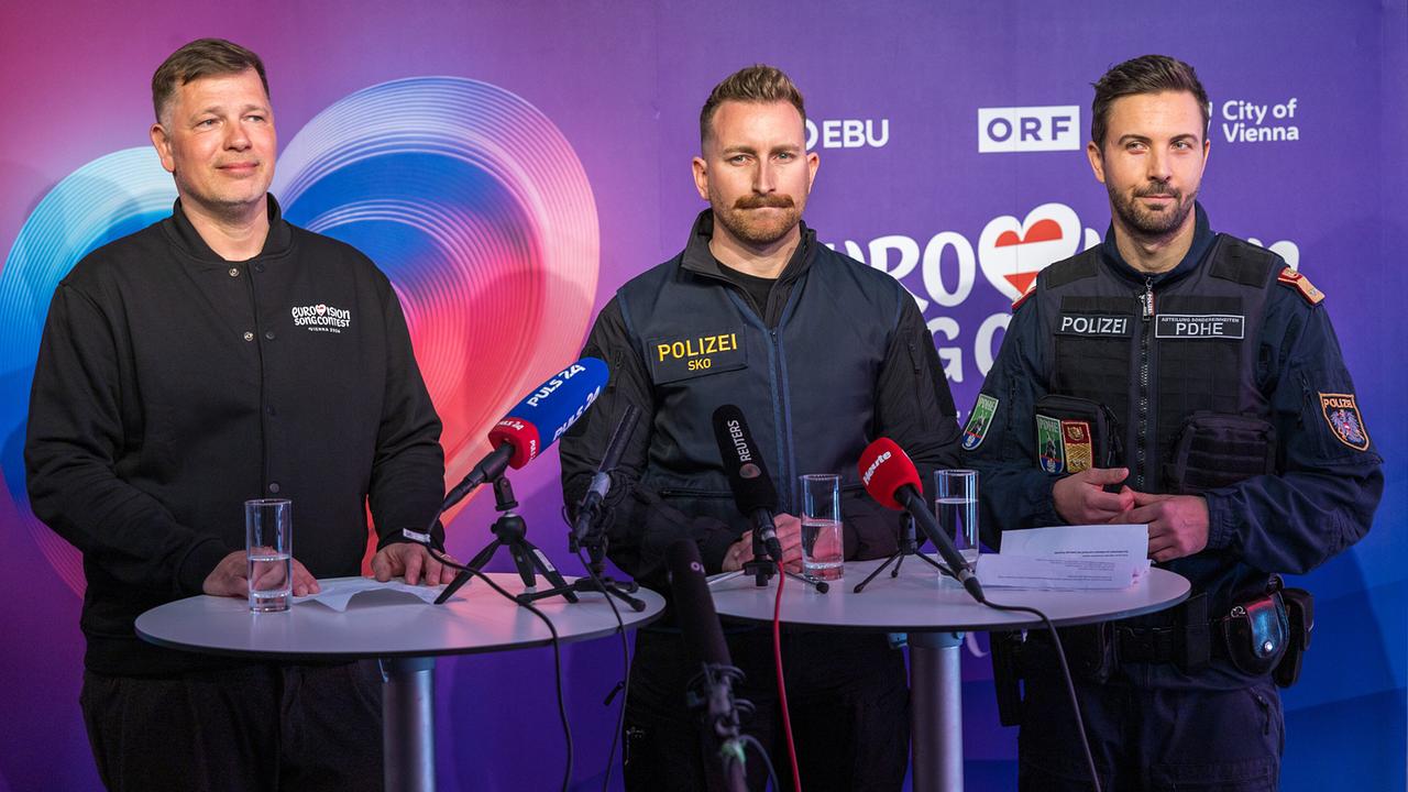 Oliver Lingens (Head of Event) and the the police officers Manuel Schneider and Gabriel Berkes stand at high tables in front of microphones during a press conference, two of them in police uniform, in front of a colored ESC background.
