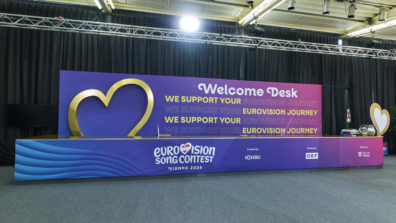 A large, colorfully designed welcome desk for the Eurovision Song Contest is set up in a hall, featuring a heart symbol, logos and the slogan "We support your Eurovision journey".
