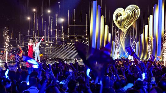 Auf der großen, hell erleuchteten Bühne stehen Victoria Swarovski und Michael Ostrowski vor jubelndem Publikum mit wehenden Fahnen, während im Hintergrund ein goldenes Herzmotiv und spektakuläre Lichteffekte zu sehen sind.