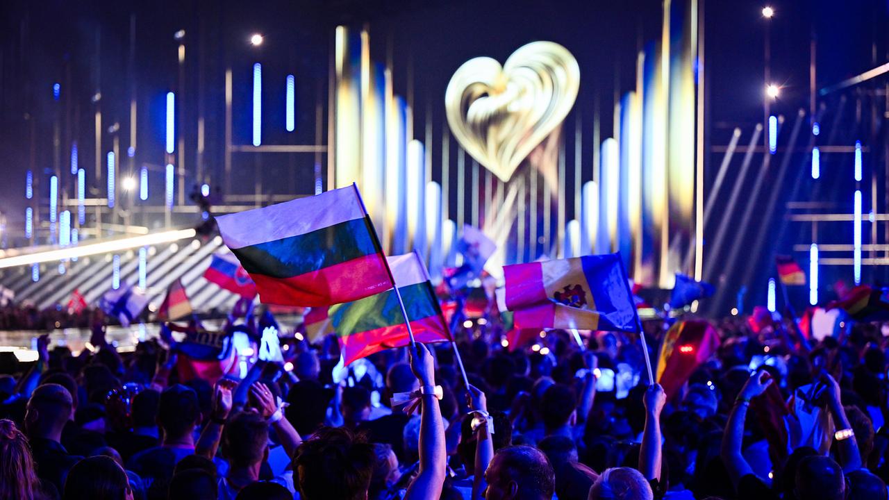 © ORF/Klaus Titzer The audience at the Vienna Stadthalle is waving flags in front of a brightly lit stage with a heart symbol at the Eurovision Song Contest 2026.