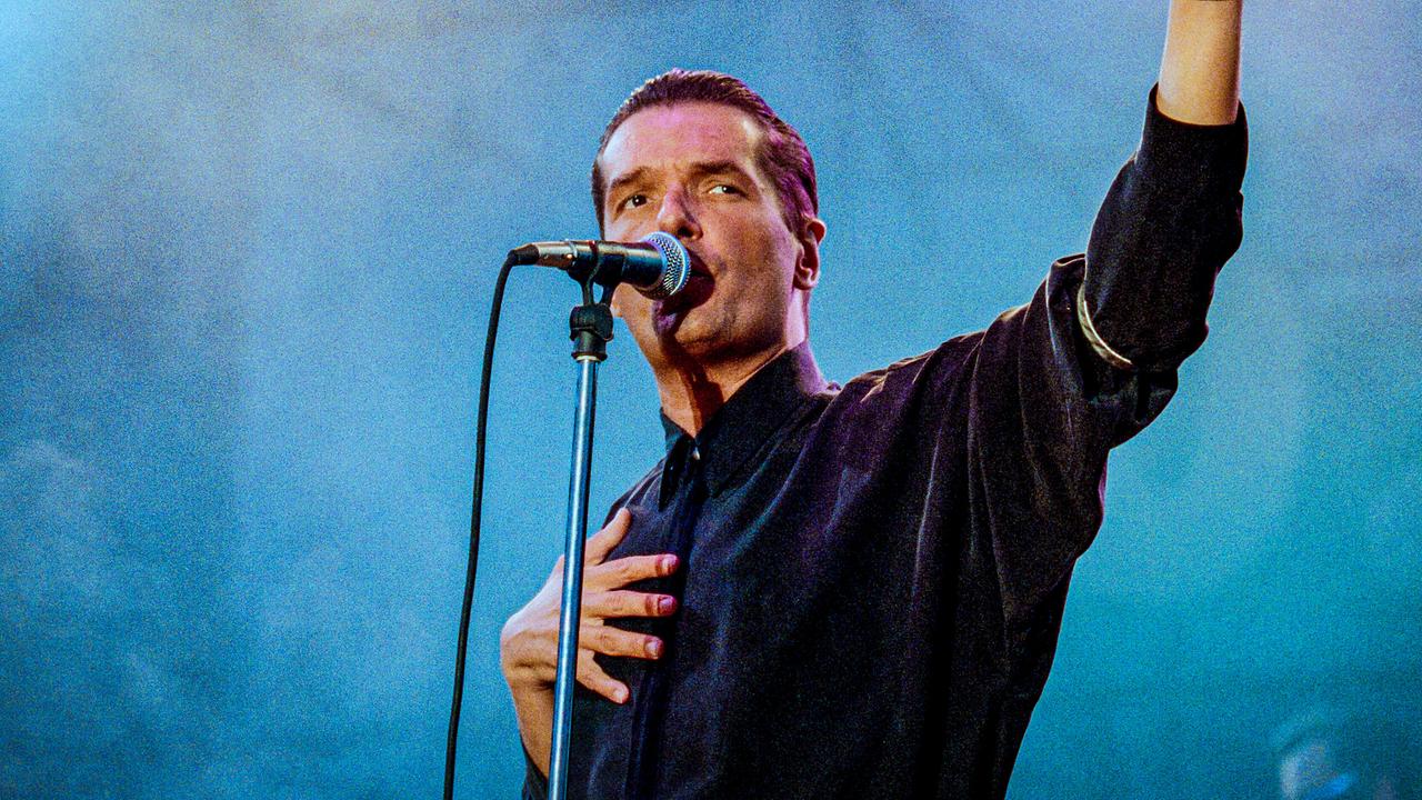 © ORF/Johannes Cizek Falco steht auf einer Bühne am Mikrofon, mit einer Hand auf der Brust und die andere nach oben gestreckt.