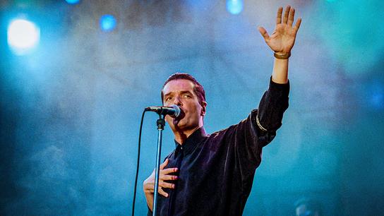 Falco steht auf einer Bühne am Mikrofon, mit einer Hand auf der Brust und die andere nach oben gestreckt.